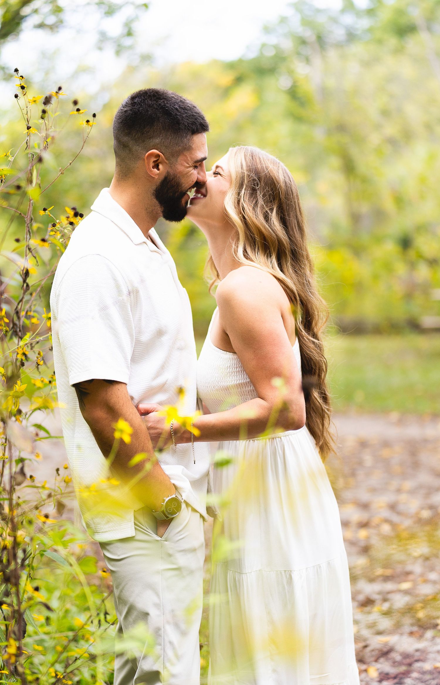 Happy couple in white outfits almost kissing surrounded by yellow wildflowers in nature.