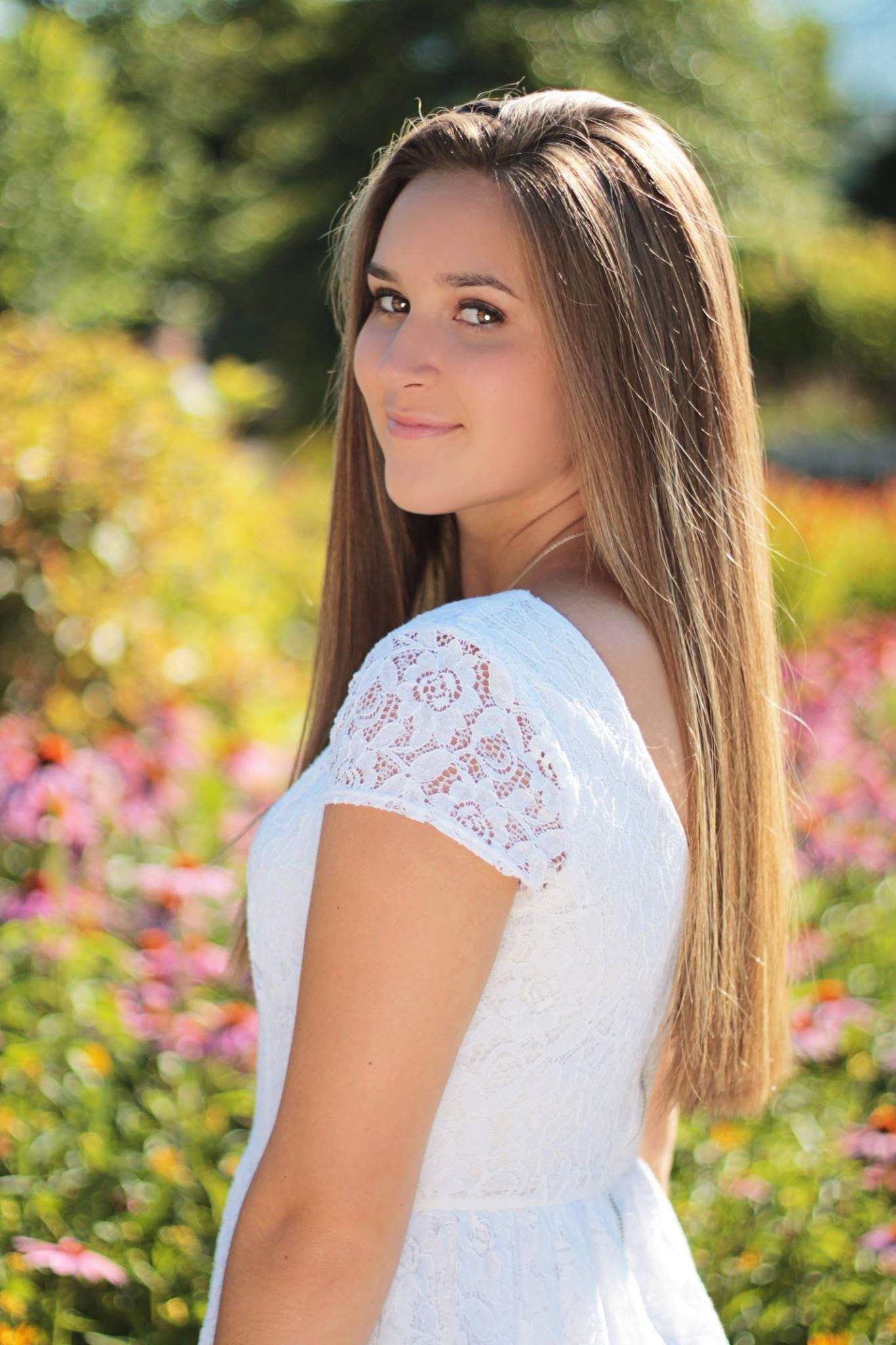 Young brunette woman in white lace dress standing in colorful flower garden on sunny day.