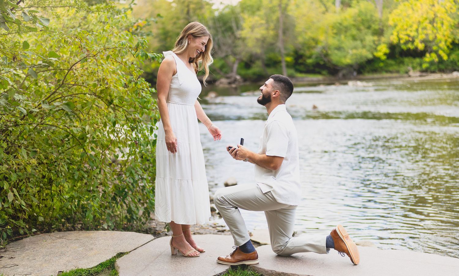 Man proposes on one knee to woman in white dress by a river surrounded by lush greenery on a sunny day.