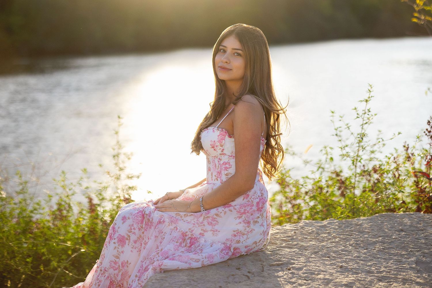 Young woman in pink floral maxi dress sitting on rock by river in warm golden sunset light.