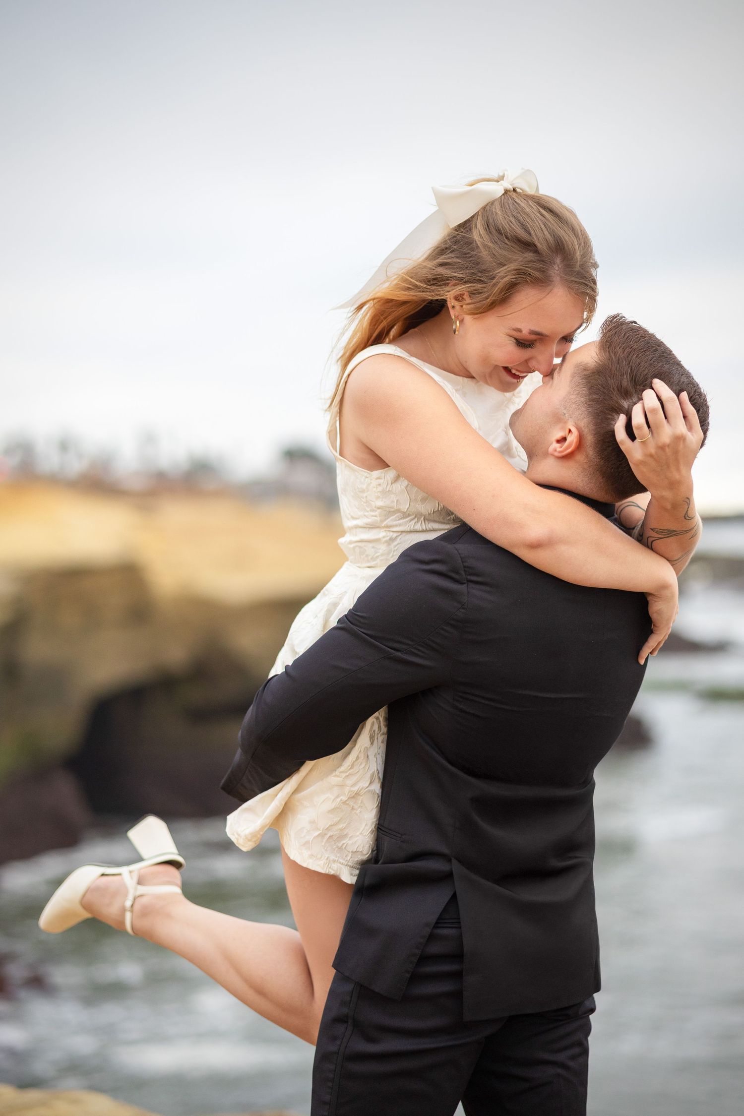 Groom lifting bride in white dress and veil on rocky ocean cliffside backdrop.
