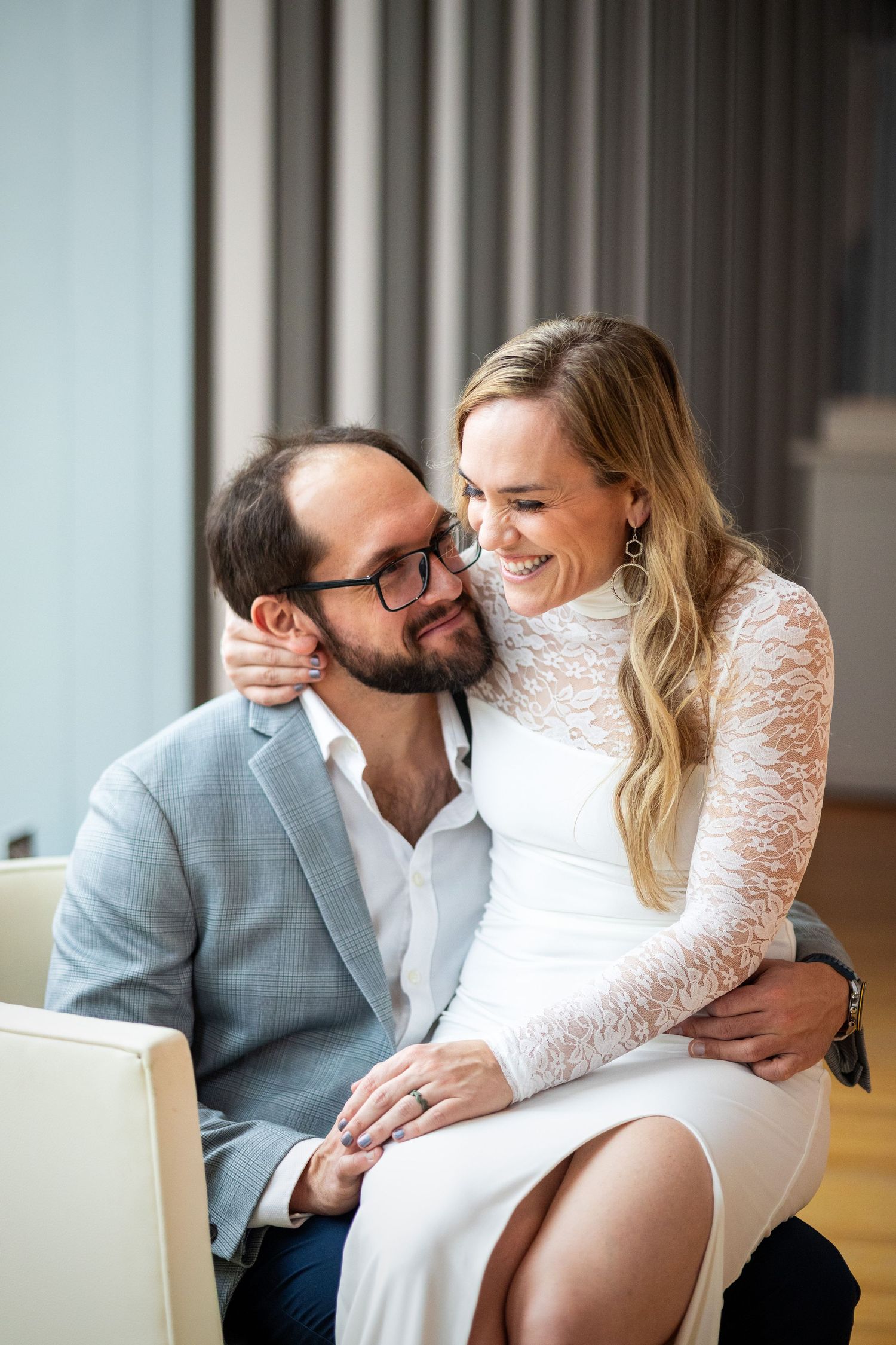Smiling couple in wedding attire embrace intimately while seated indoors in soft light.