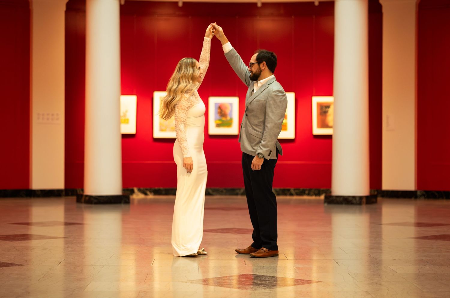 Couple dancing together in a grand red-walled art museum hall, woman in white jumpsuit and man in gray suit.
