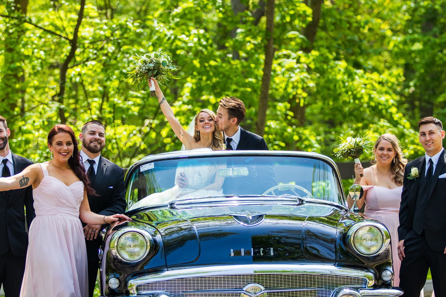 Bride and groom kiss atop a classic vintage black car surrounded by their wedding party in a lush green setting.