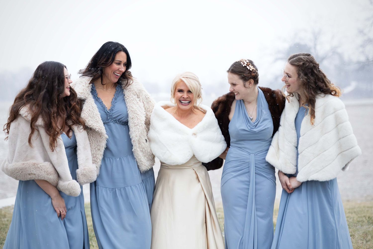 Smiling bride in ivory fur shawl surrounded by four bridesmaids in light blue gowns and faux fur wraps outdoors.