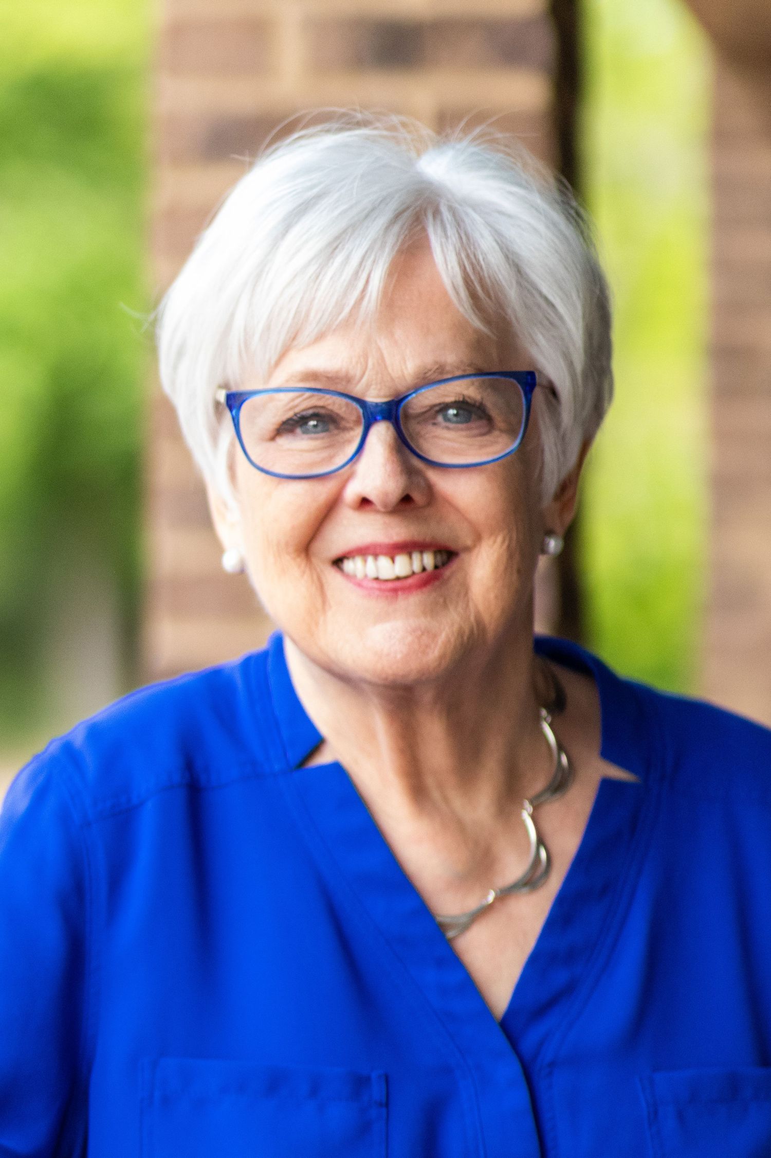 Smiling senior woman with white hair and blue glasses wearing a blue blouse for a professional headshot.