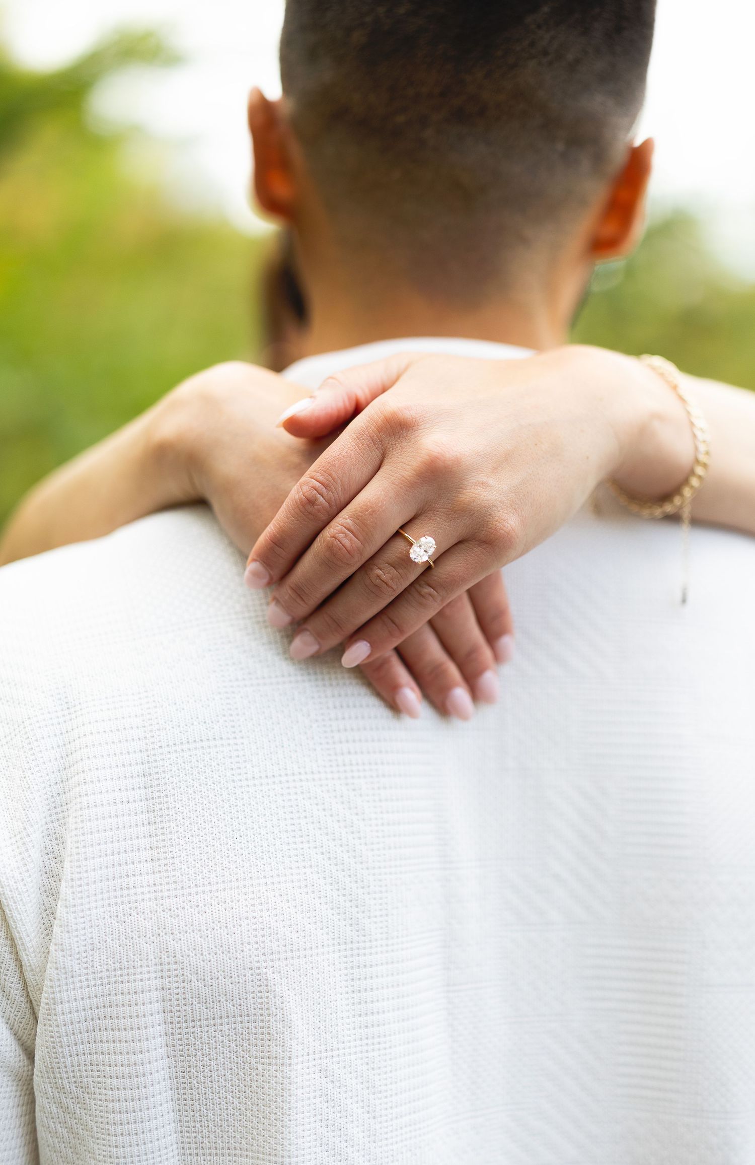 A woman's hand with an engagement ring rests on a man's shoulder, both dressed in white outdoors.