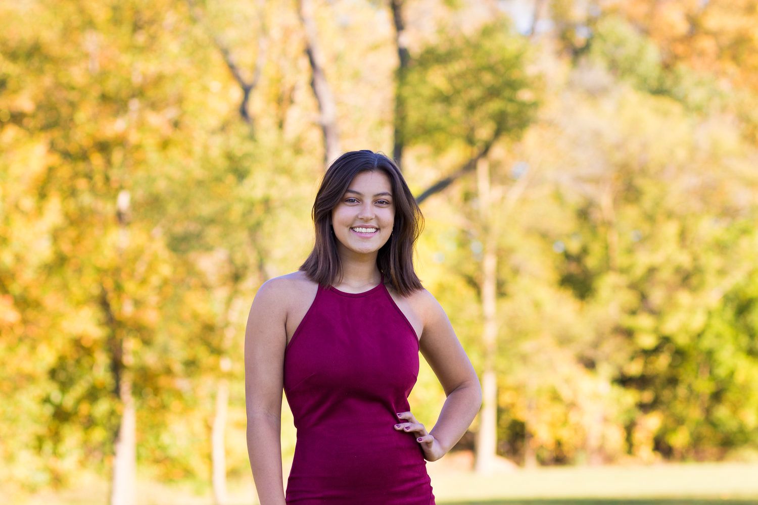 Smiling young woman in magenta dress posing outdoors with colorful autumn foliage background.