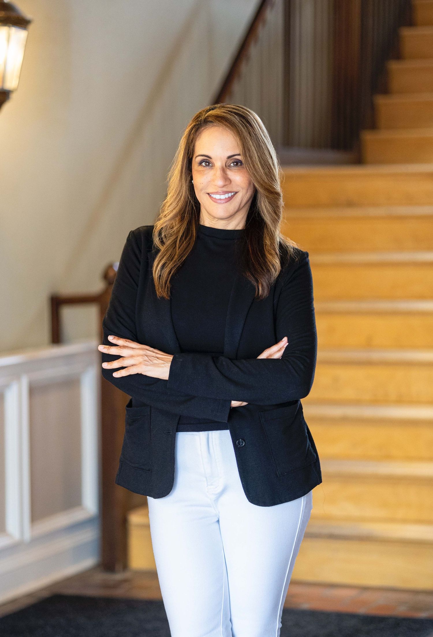 Confident woman in black blazer and white pants standing on elegant staircase with arms crossed.