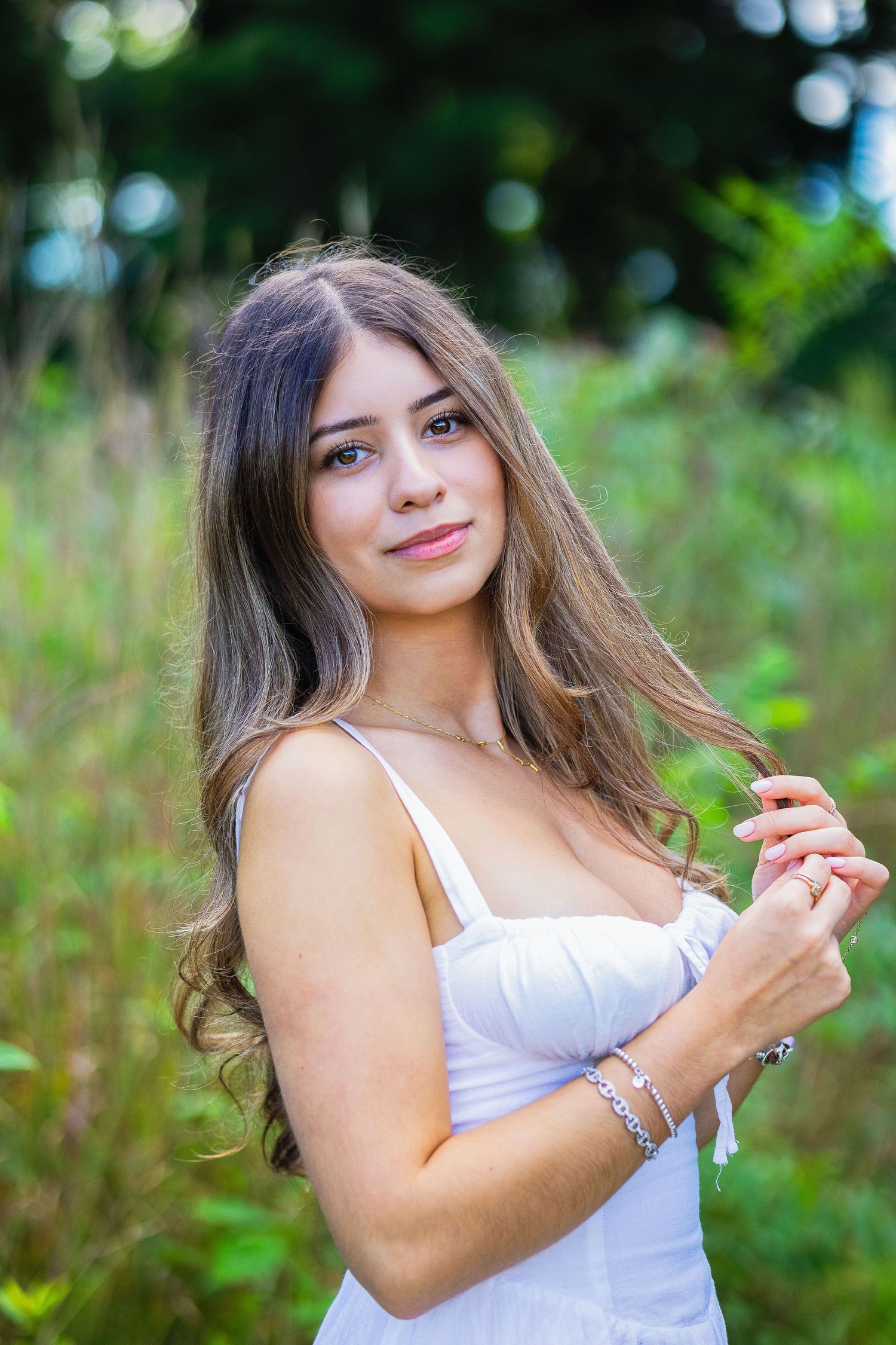 Young woman with long brown hair in white dress smiling outdoors in green nature setting.