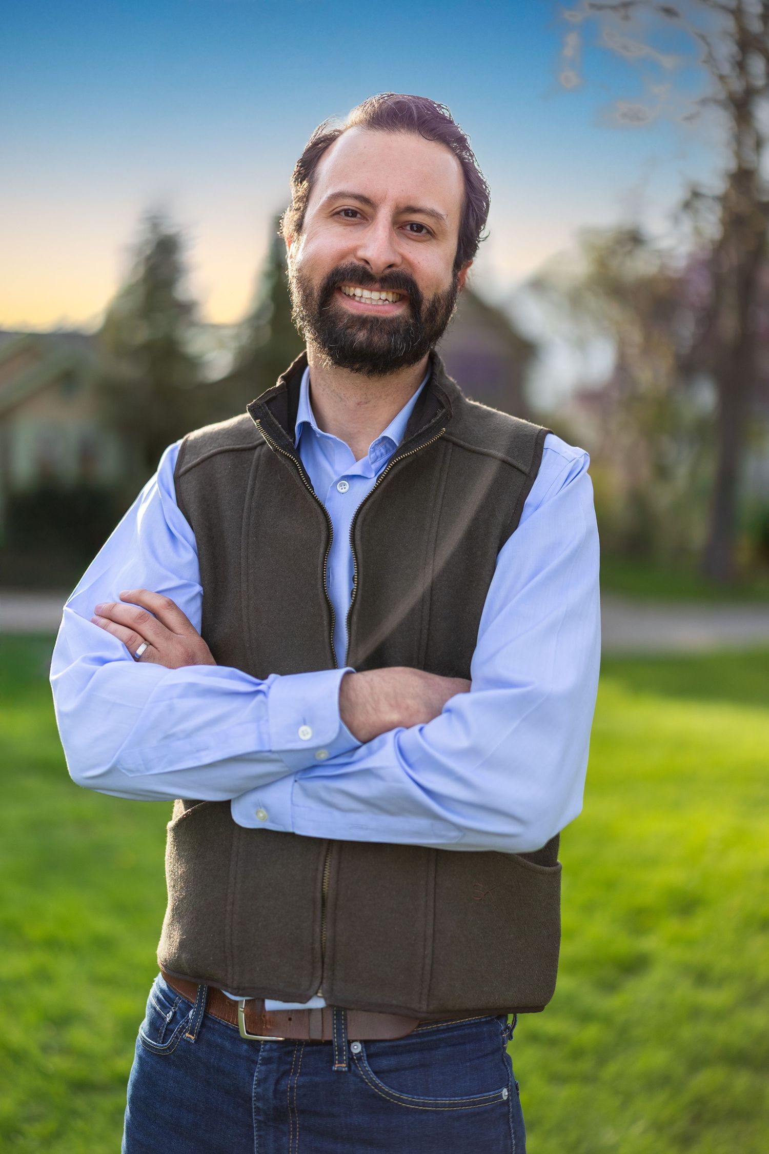 Bearded man in vest and blue shirt standing outdoors with arms crossed.