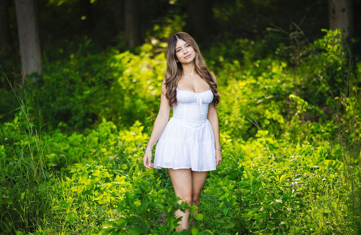 Smiling young woman in white mini dress strolling through vibrant green forest in soft sunlight.
