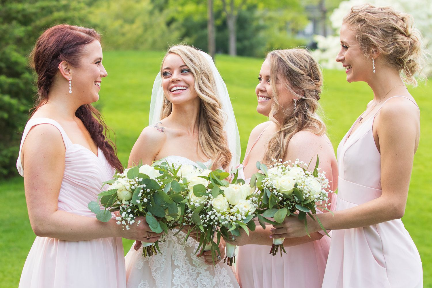 Bride in lace gown smiles with three bridesmaids in blush pink dresses holding white and greenery bouquets outdoors.