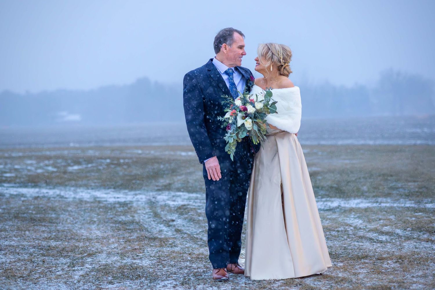 A bride and groom stand together in a snowy open field, the bride holding a colorful bouquet on their winter wedding day.