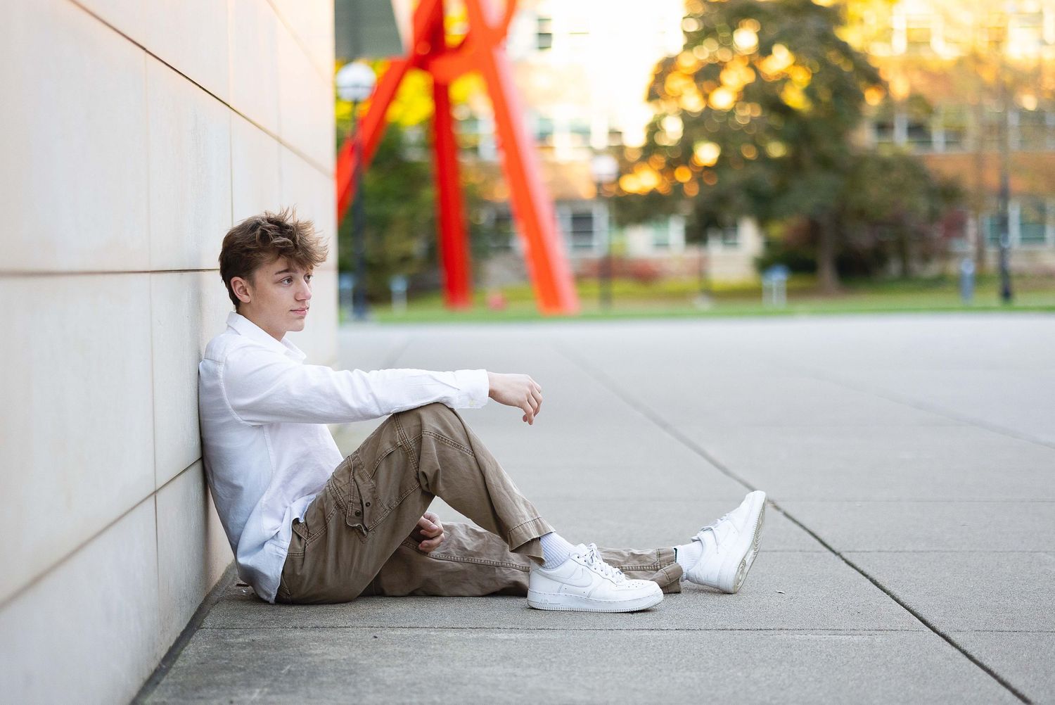 Young man in white shirt and khaki pants sitting against a wall outdoors at golden hour.