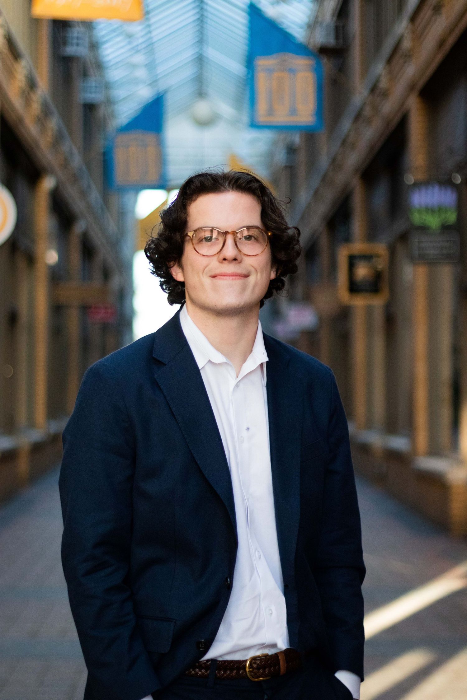 Young man in navy blazer and glasses smiling in elegant indoor shopping arcade hallway.