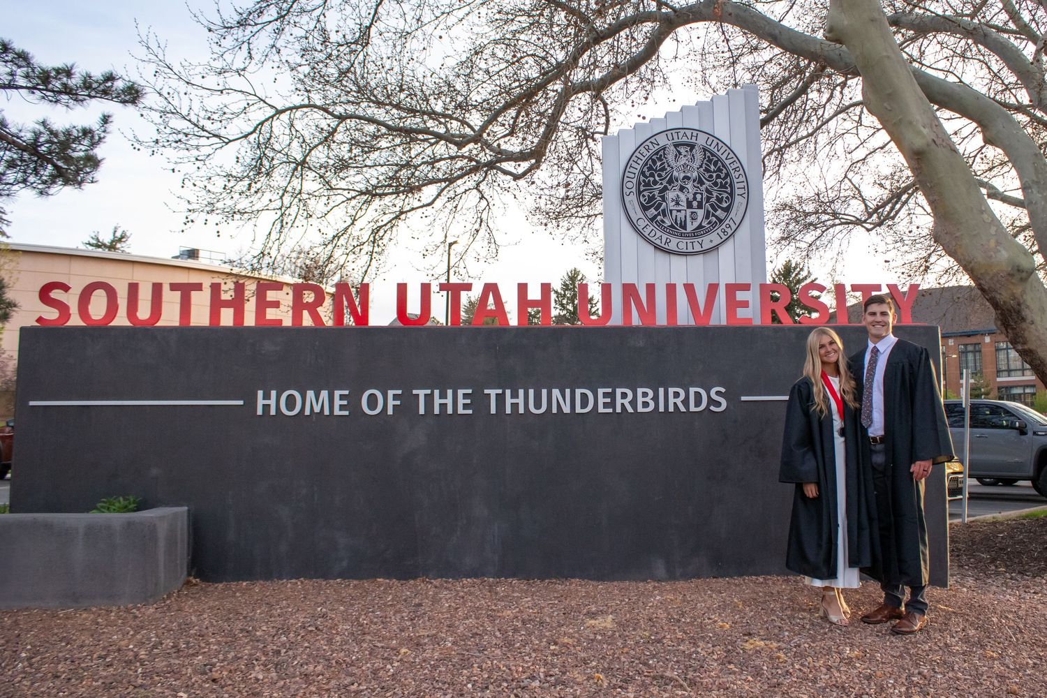 Two graduates pose proudly in front of Southern Utah University's iconic sign, Home of the Thunderbirds.