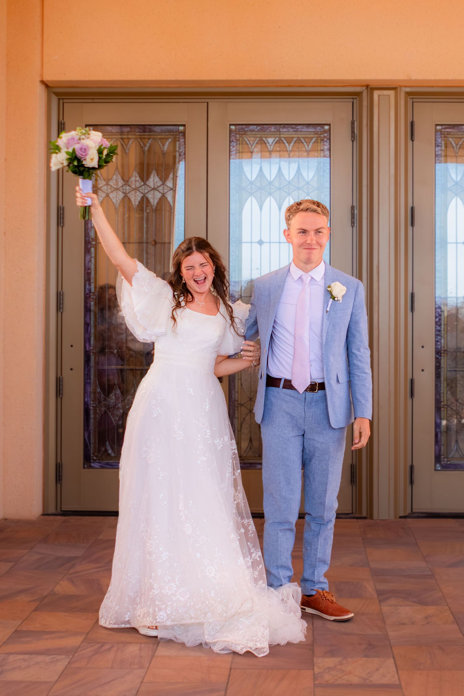 Joyful LDS bride in white lace gown raises bouquet outside doors at the Red Cliff Utah Temple in St. George Utah