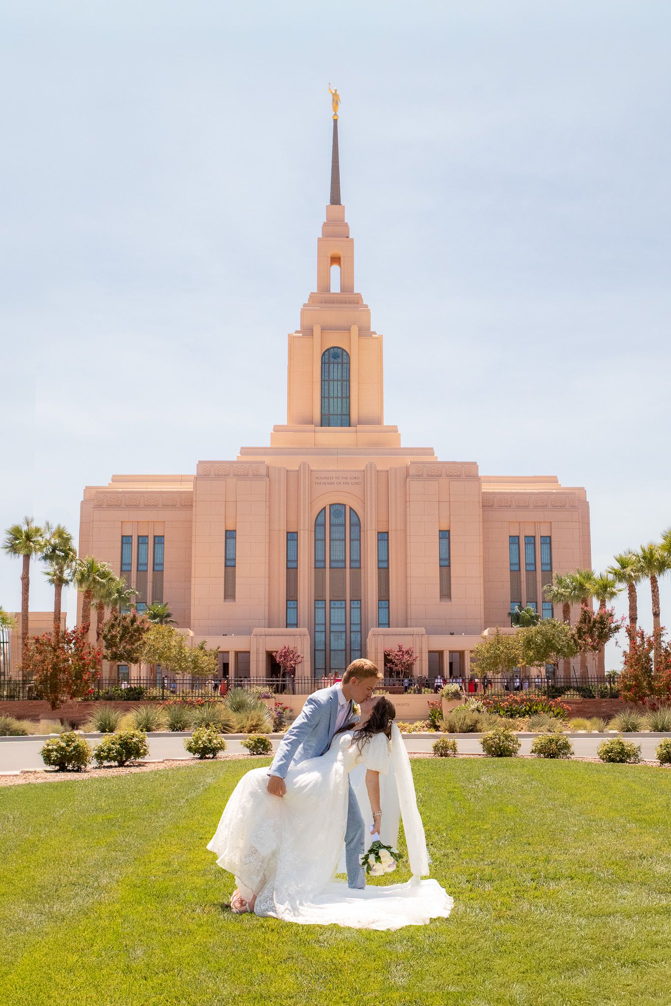 Bride and groom sharing a romantic dip kiss on green lawn in front of a grand LDS temple with a golden spire.