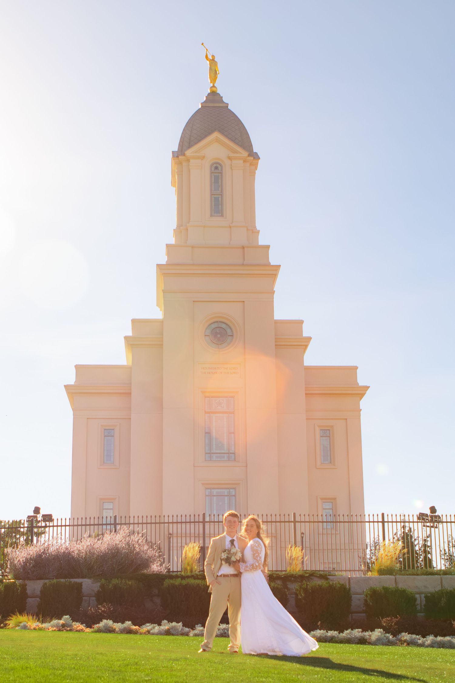 Bride and groom pose in front of a sunlit LDS temple with golden angel statue atop the steeple.