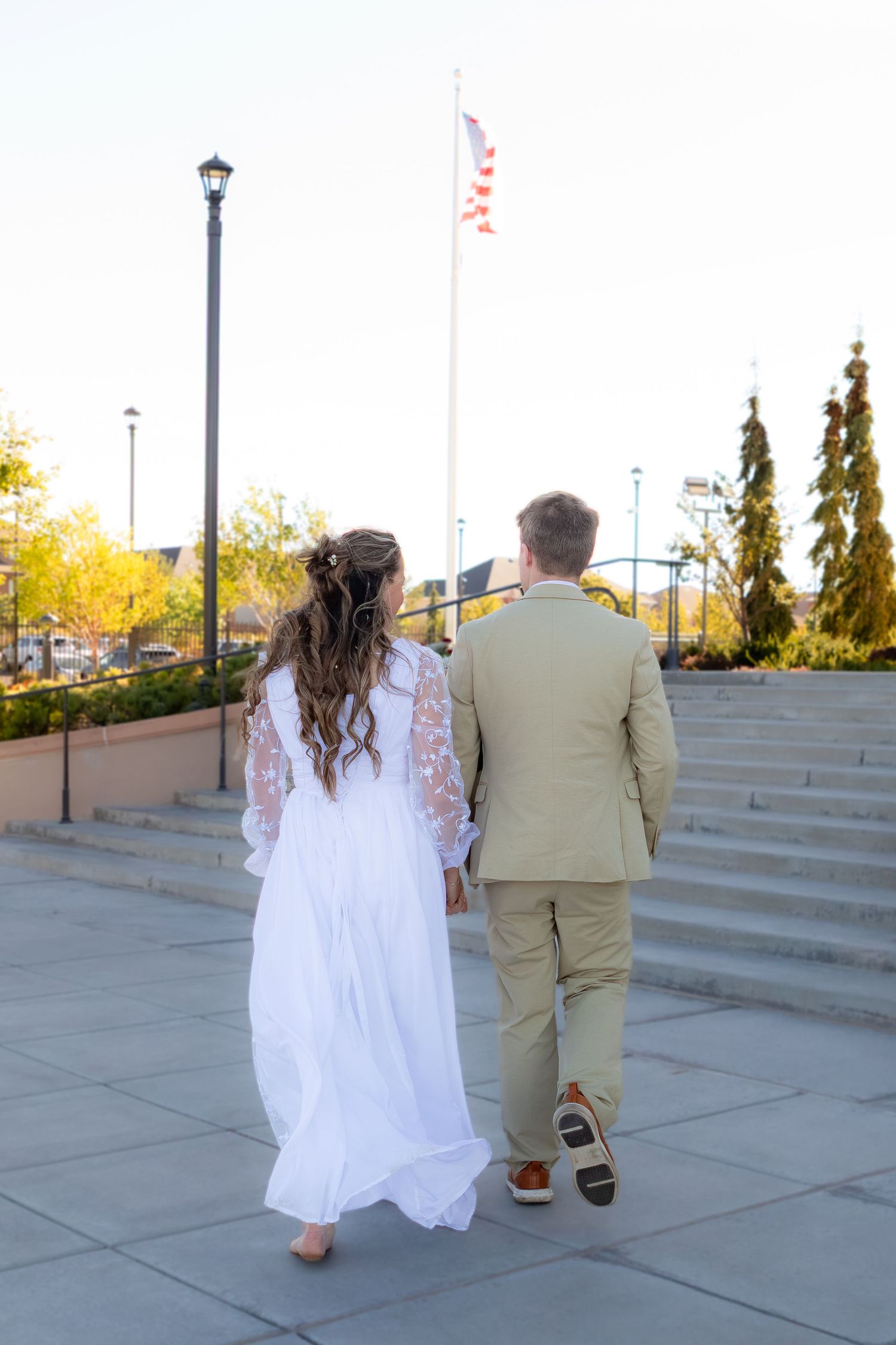 Bride and groom walking hand in hand toward steps near an American flag on their wedding day.