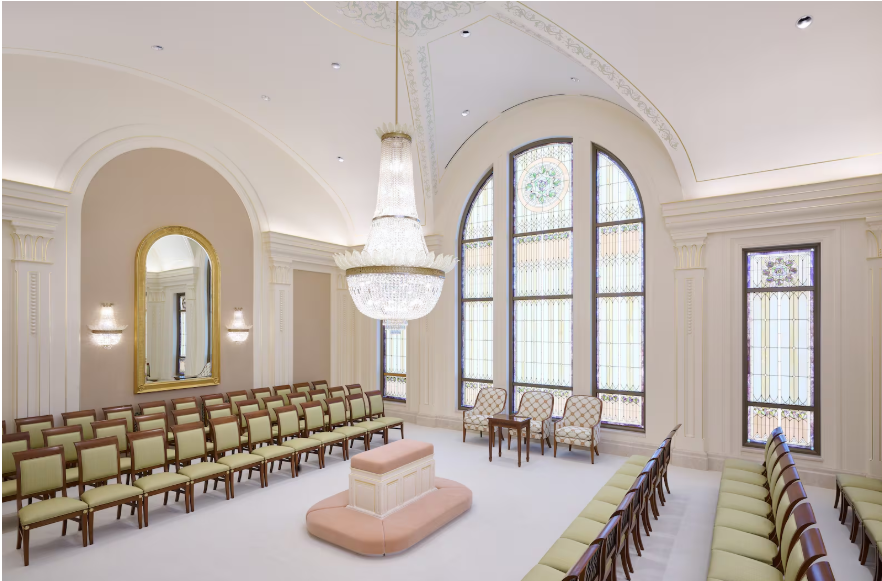 Elegant LDS temple celestial room with crystal chandelier, arched stained glass windows, white walls, and rows of gold chairs.
