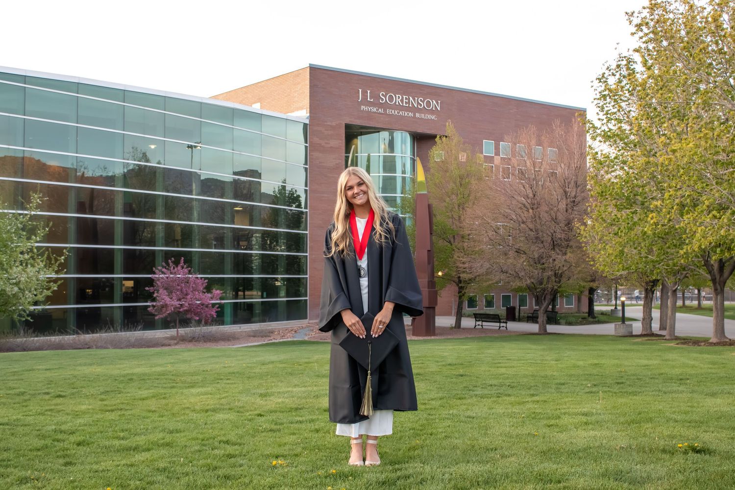 A young woman in graduation cap and gown stands proudly outside the J.L. Robinson School of Business building on a sunny day.