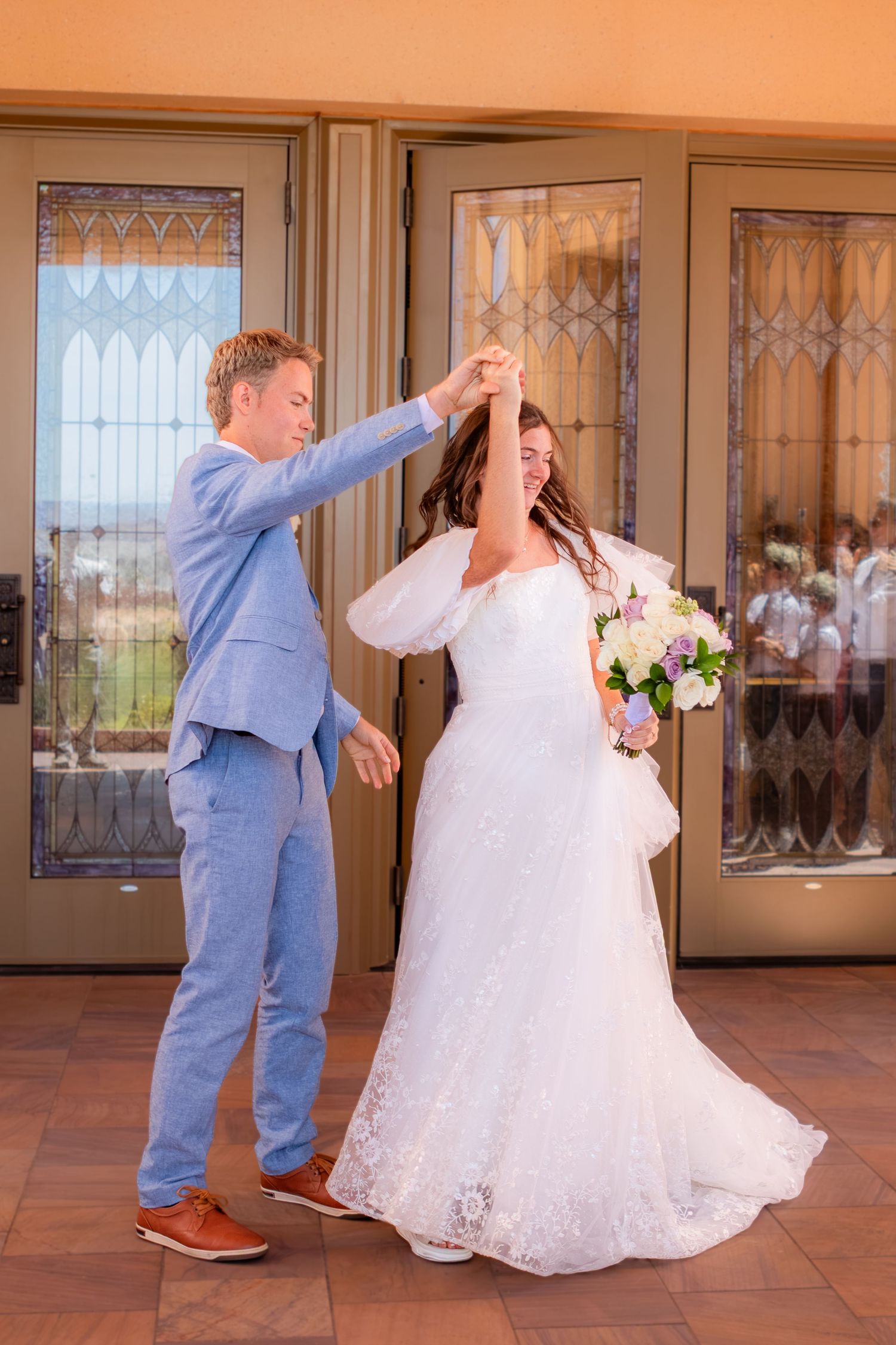 LDS Groom in blue suit twirls bride in white lace wedding gown holding floral bouquet outside doors. Red Cliffs Temple Saint George Utah