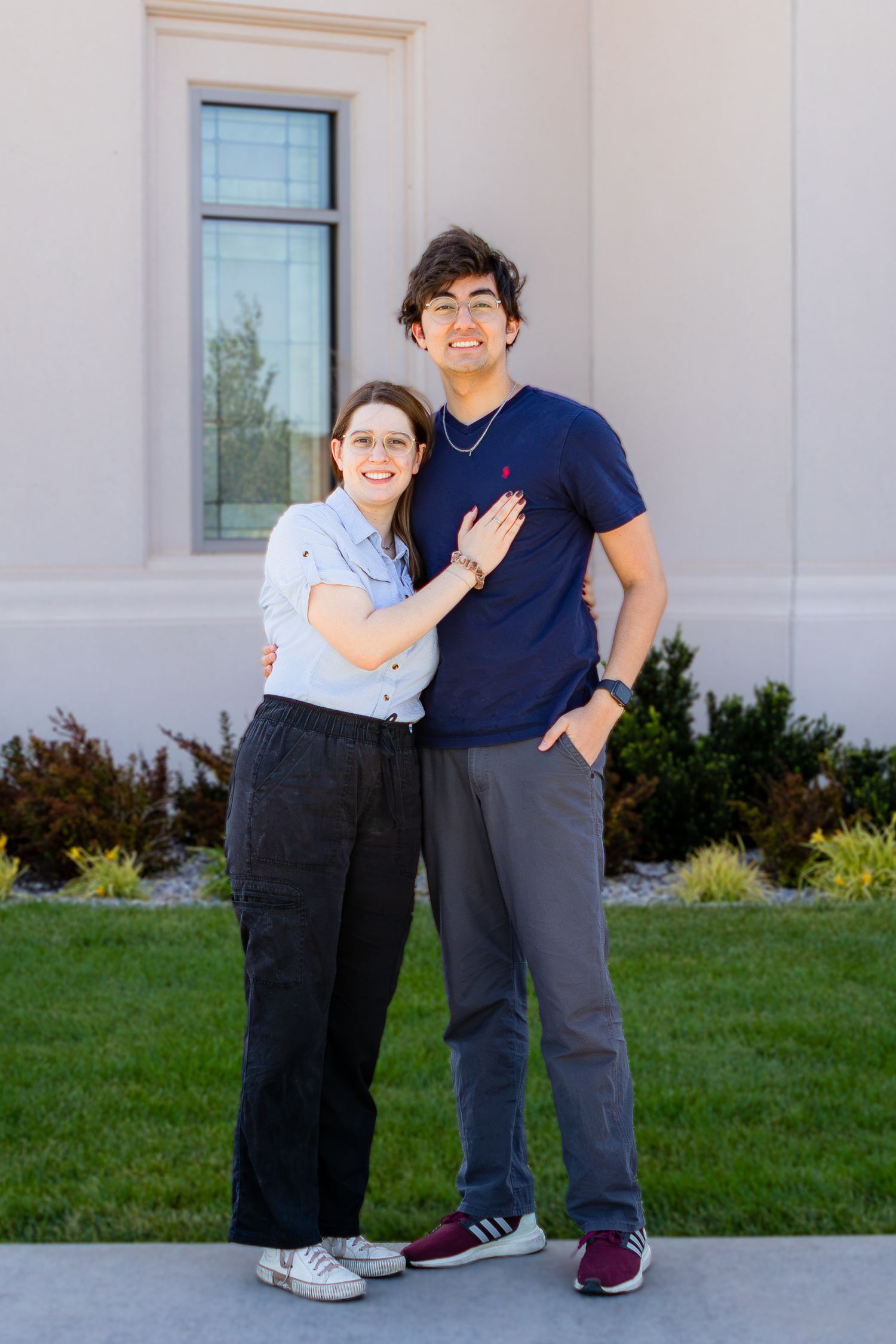A smiling young couple poses together outdoors in front of a building, dressed casually in blue and gray outfits.