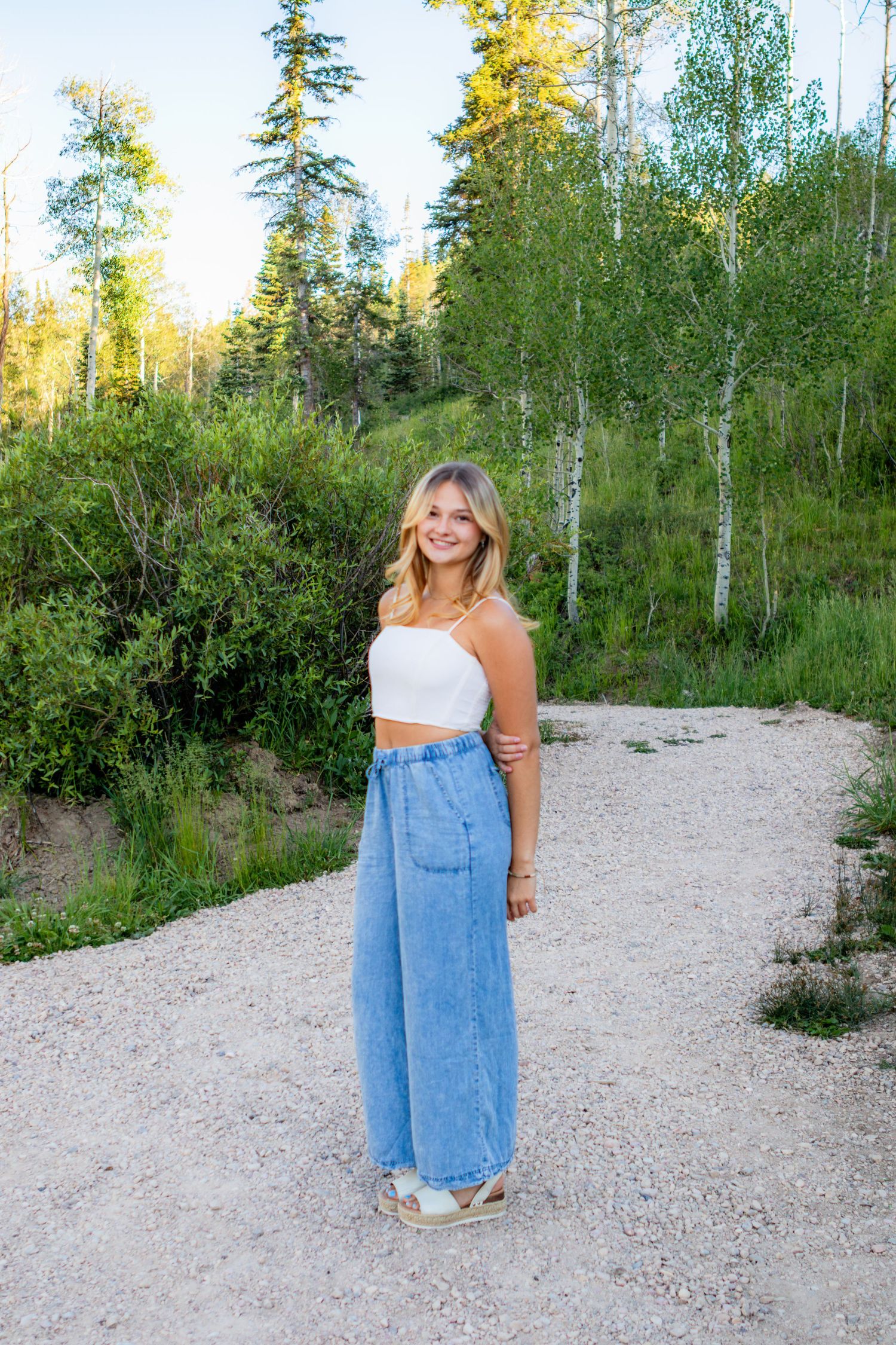 Young blonde woman smiling in white crop top and wide-leg jeans on a gravel path surrounded by lush green trees.