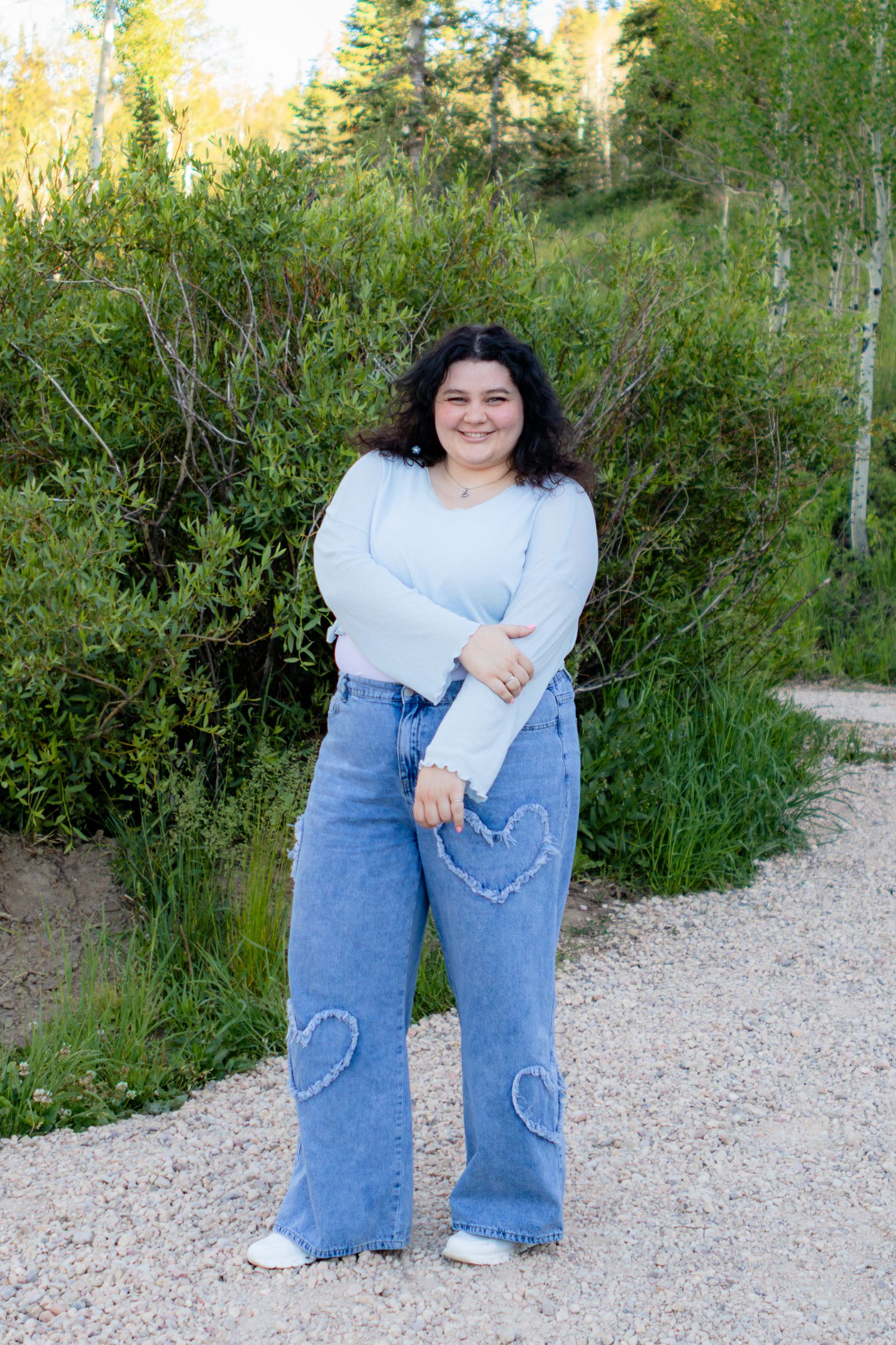 Young woman in wide-leg heart-patch jeans and blue top smiling on a gravel path surrounded by green trees.