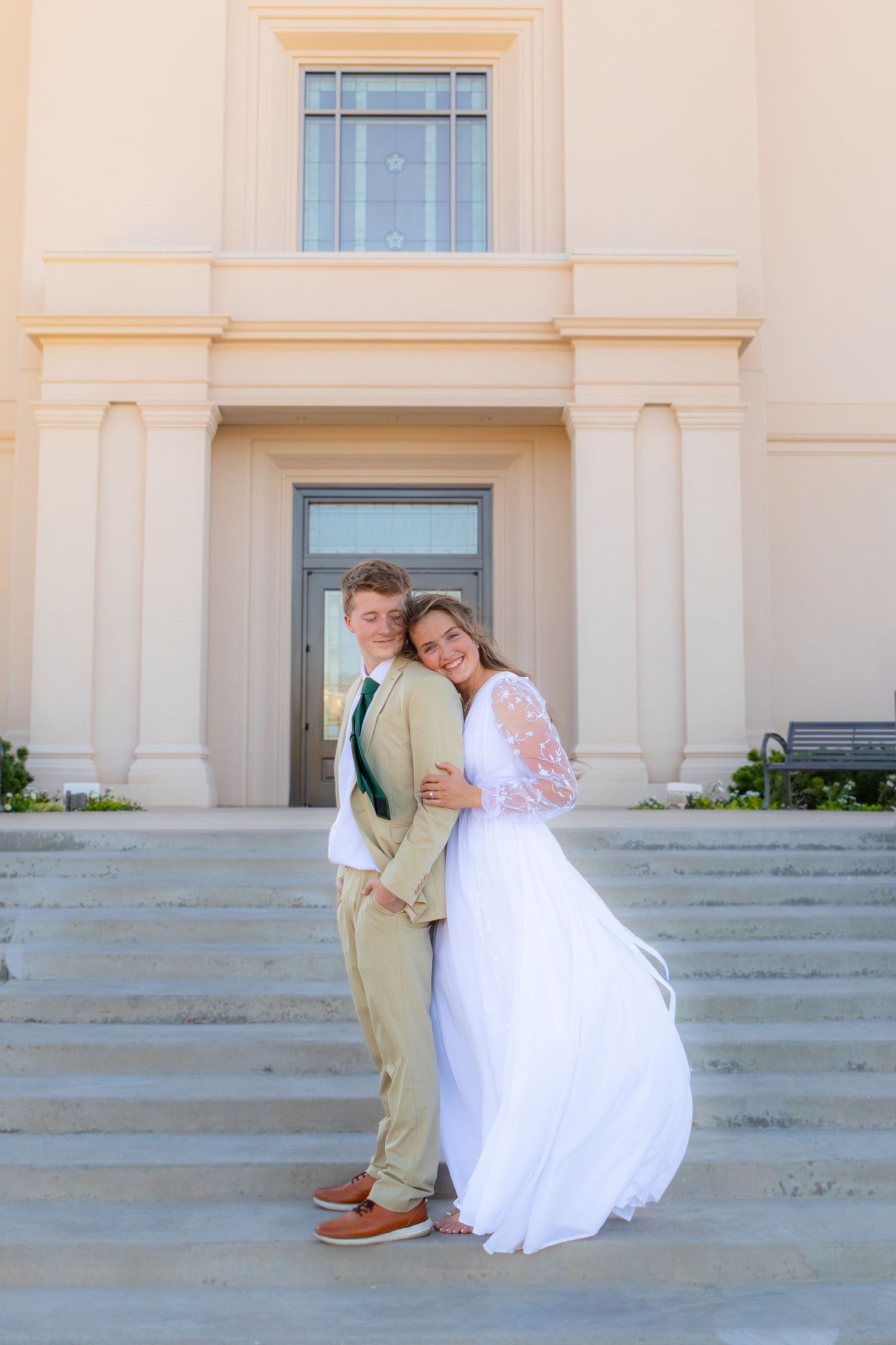 Young couple in formal attire embracing on steps of elegant building, groom in tan suit, bride in white gown.