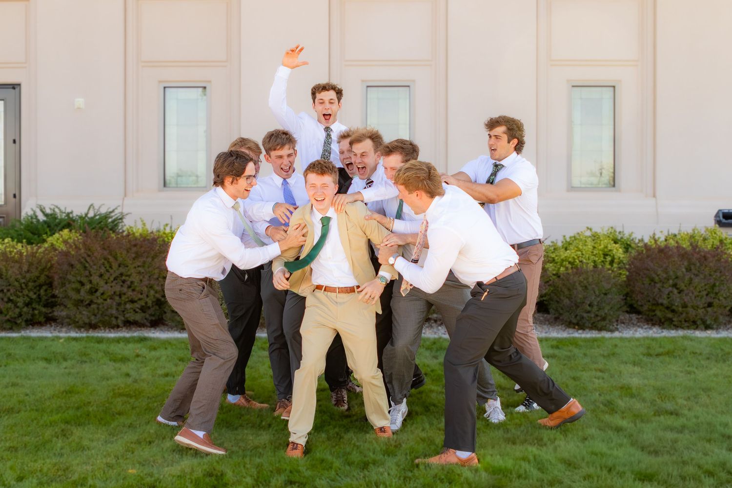 Groom in tan suit surrounded by groomsmen in white shirts playfully celebrating outdoors before wedding ceremony.