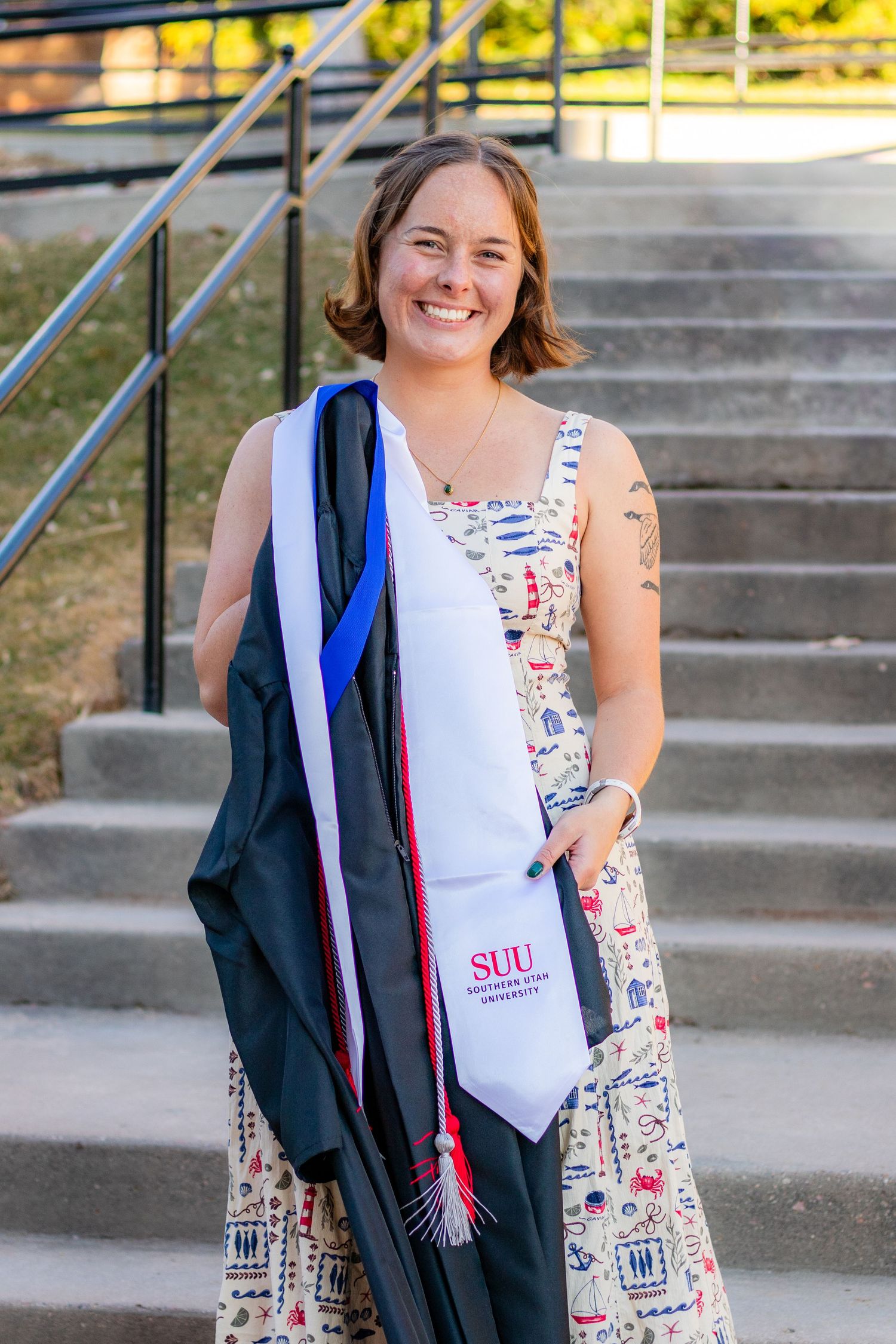 Smiling graduate holding cap and gown with SUU stole on steps outdoors.