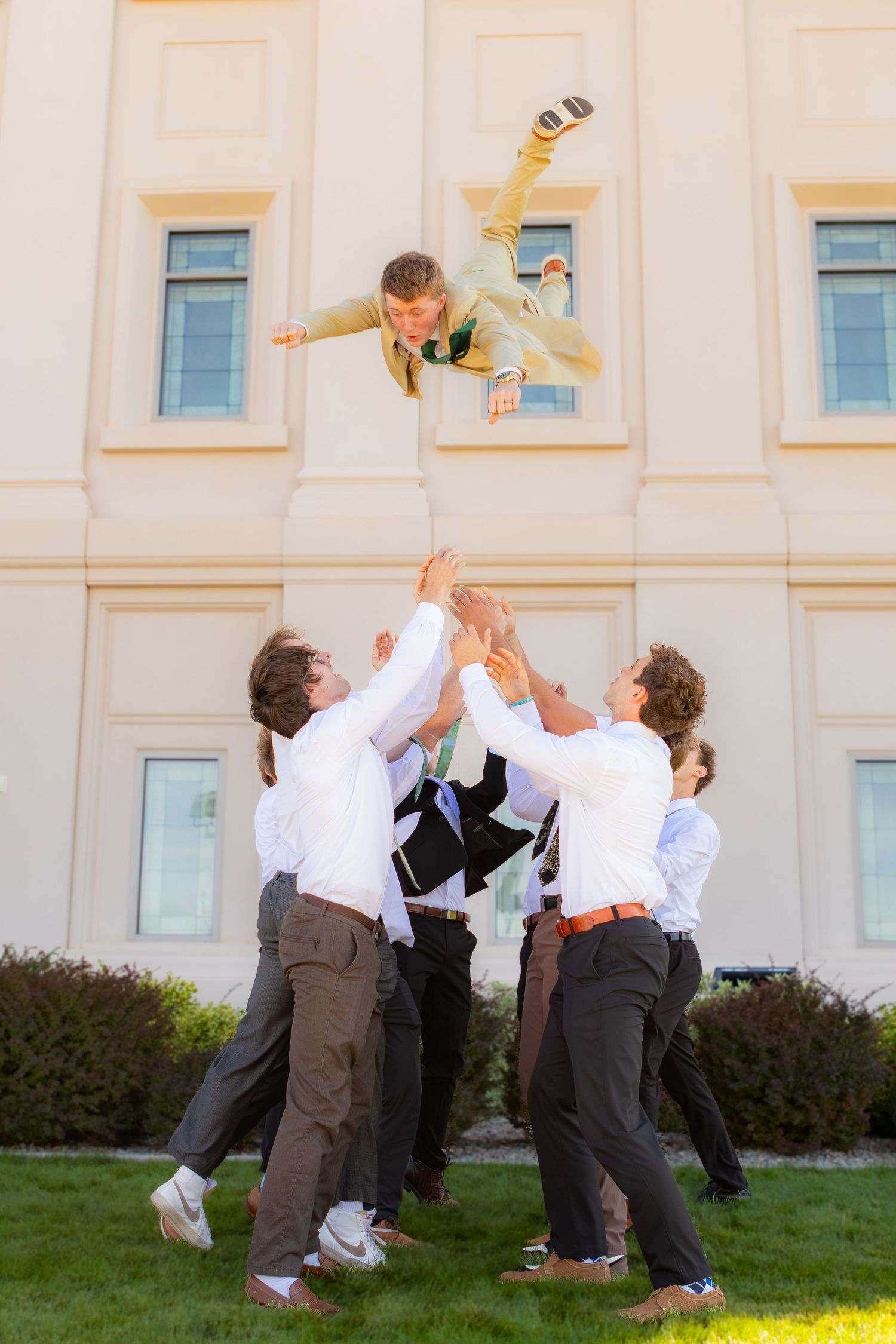 Groomsmen in white shirts tossing a bride into the air outdoors near a large building on a sunny day.