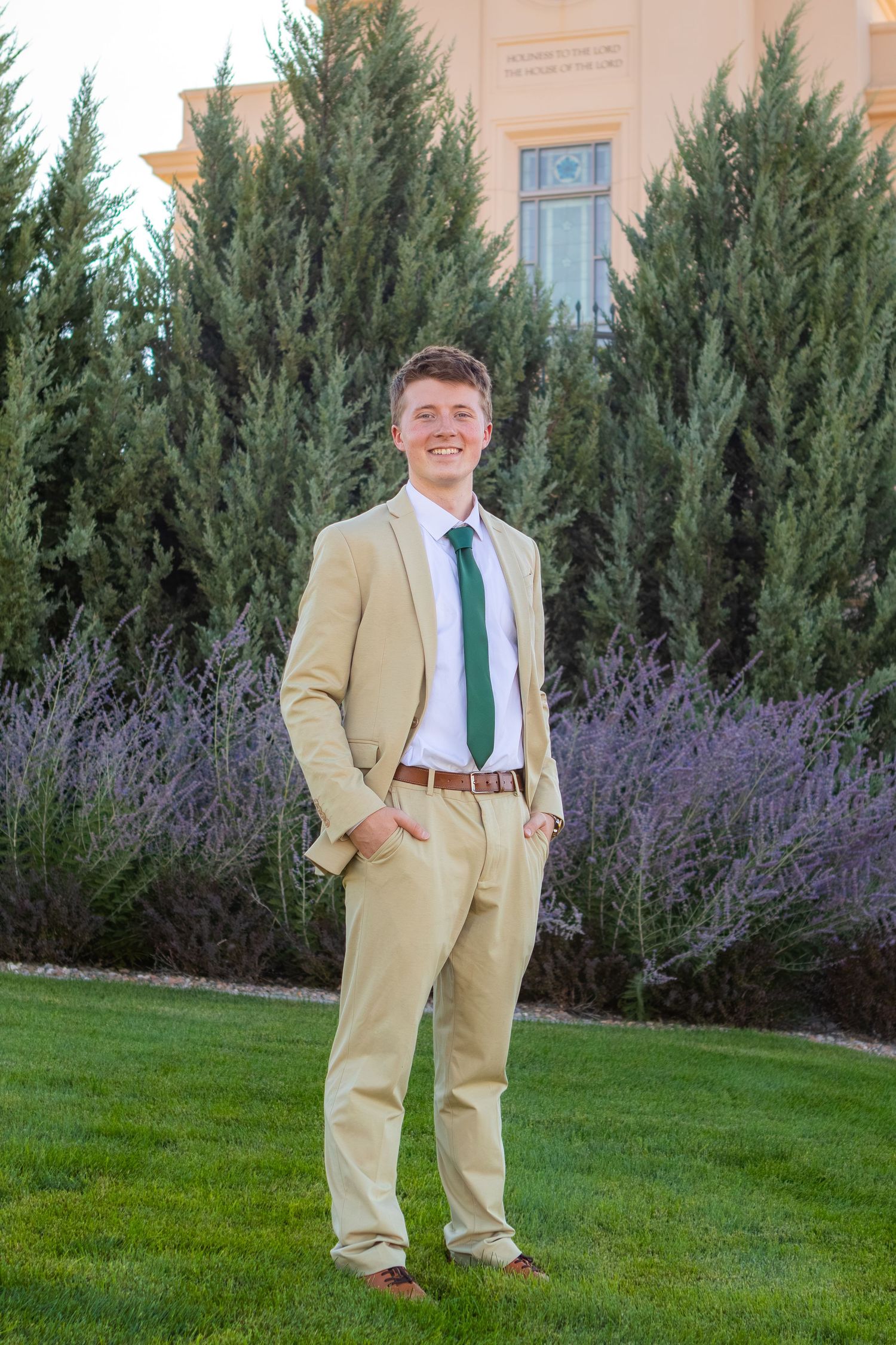 Young man in tan suit with green tie smiling outdoors on green lawn with evergreen trees and lavender bushes.