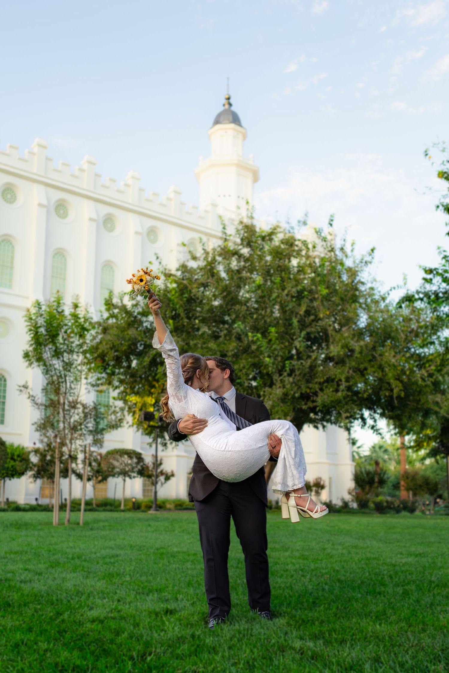 Groom lifts bride in white dress outside St. George LDS temple on green lawn, sharing a kiss while she holds a bouquet in Utah.