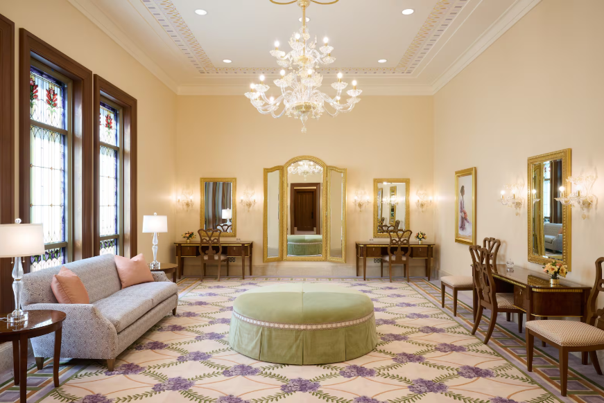 Elegant hotel lobby with chandelier, ornate mirrors, floral carpet, green ottoman, sofa, and gold accents in warm cream tones.
