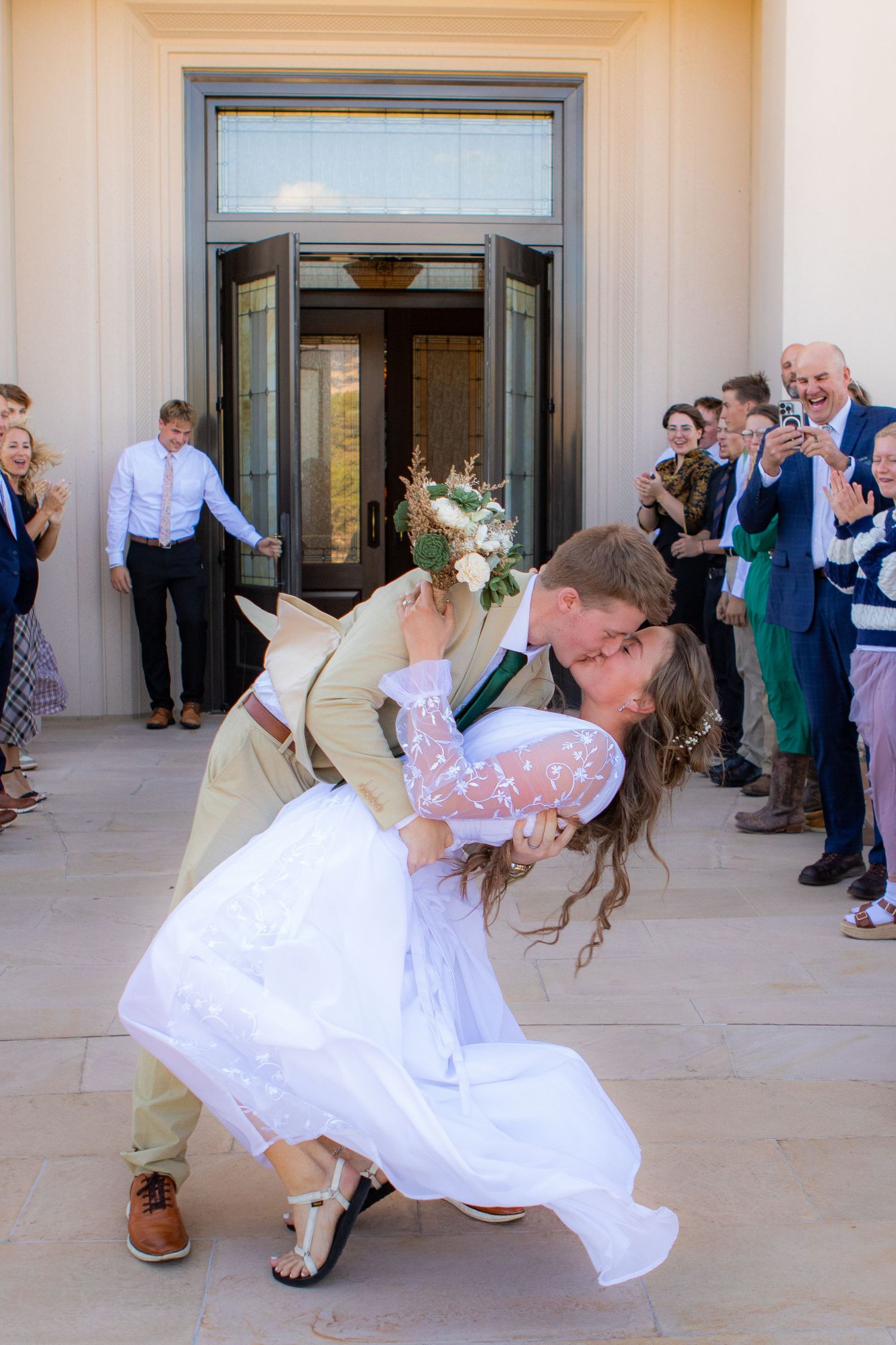 Groom dips bride for a kiss outside wedding venue as guests cheer and celebrate the newlyweds.