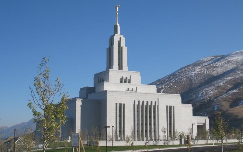 White LDS Mormon temple with golden spire against blue sky and snow-capped mountains in background.