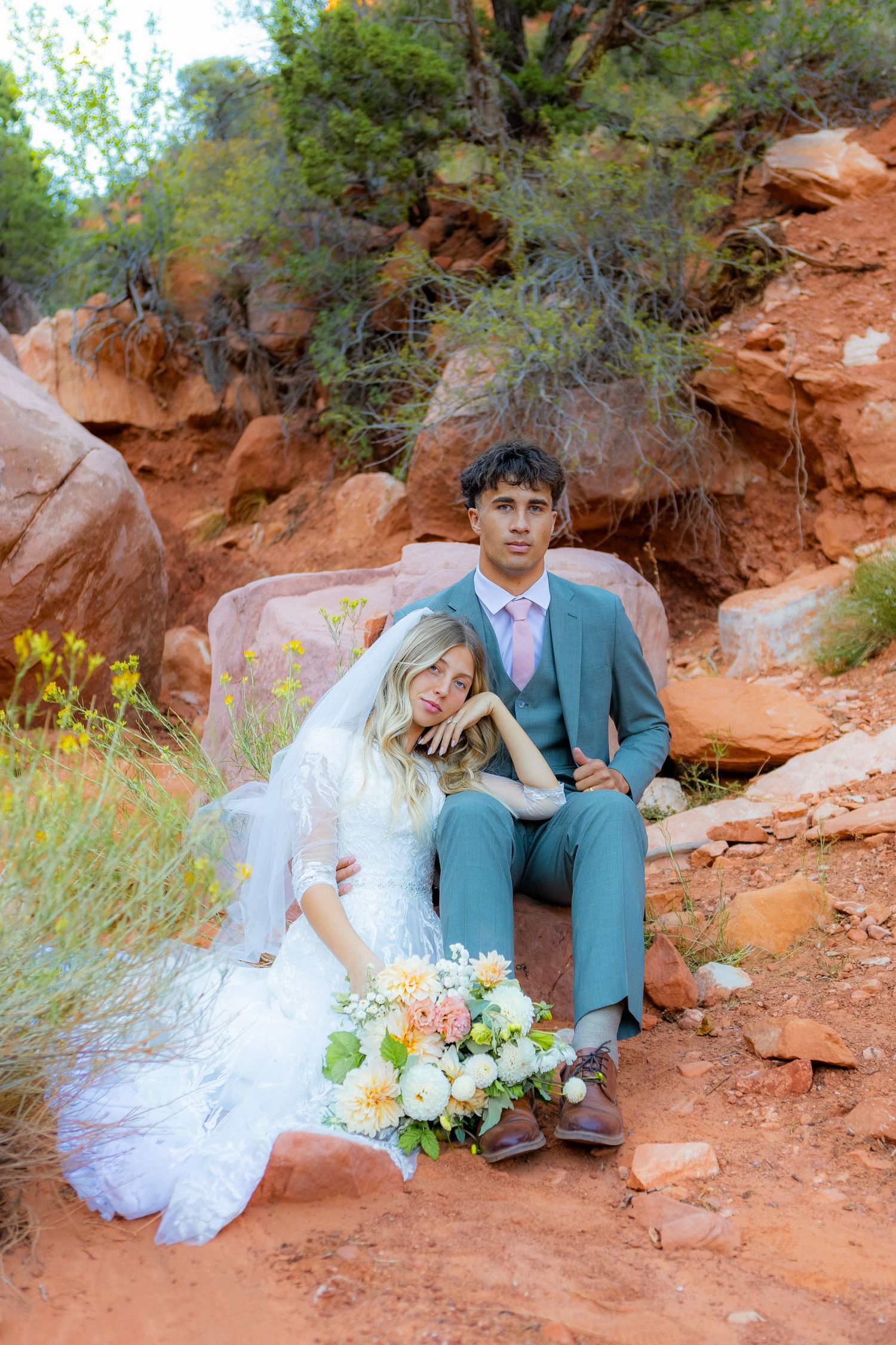 Bride and groom pose on red rocks in Sedona, Arizona, surrounded by desert flora in wedding attire.