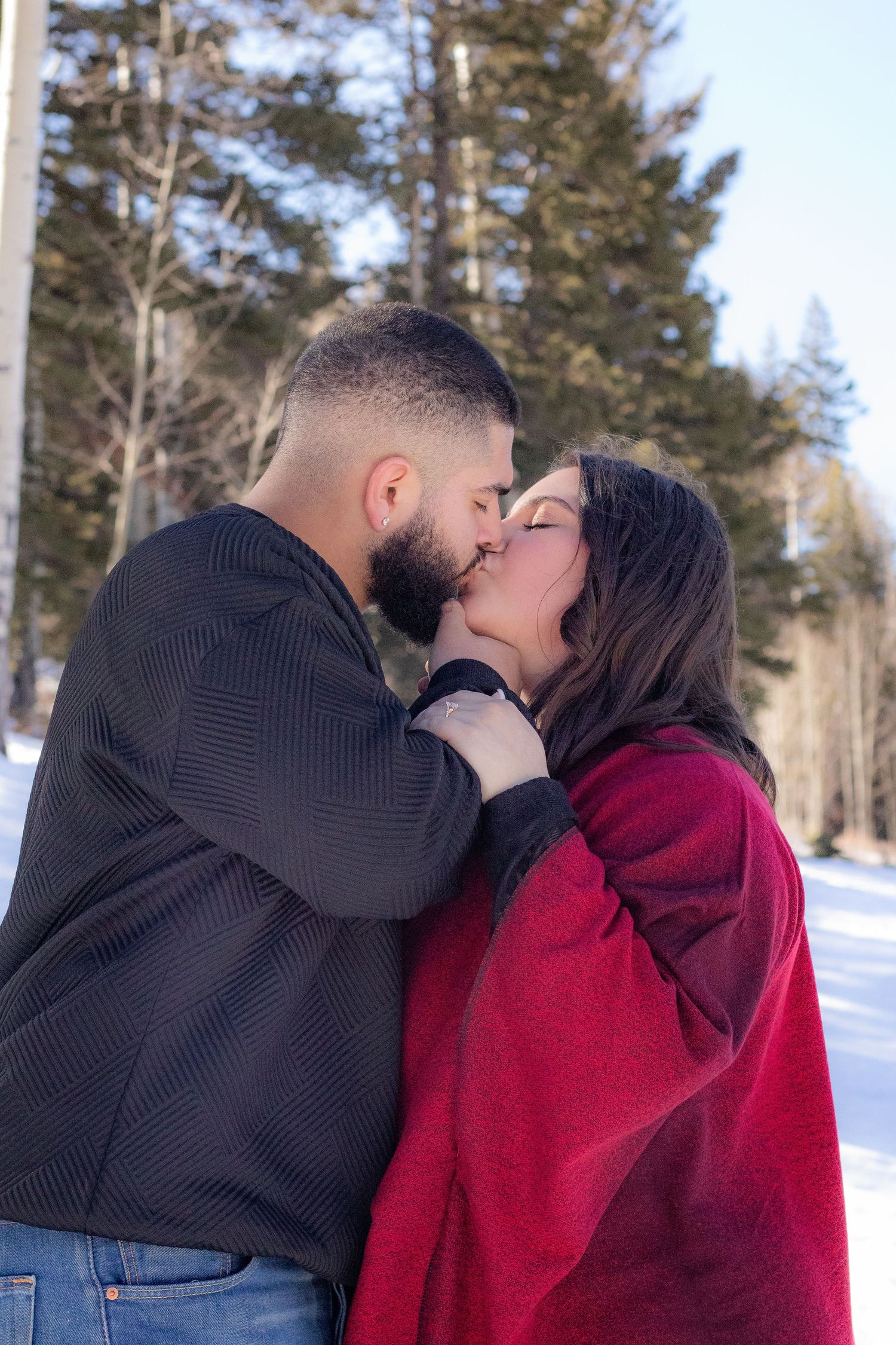 Couple shares a romantic kiss outdoors in a snowy winter forest setting wrapped in red blanket.