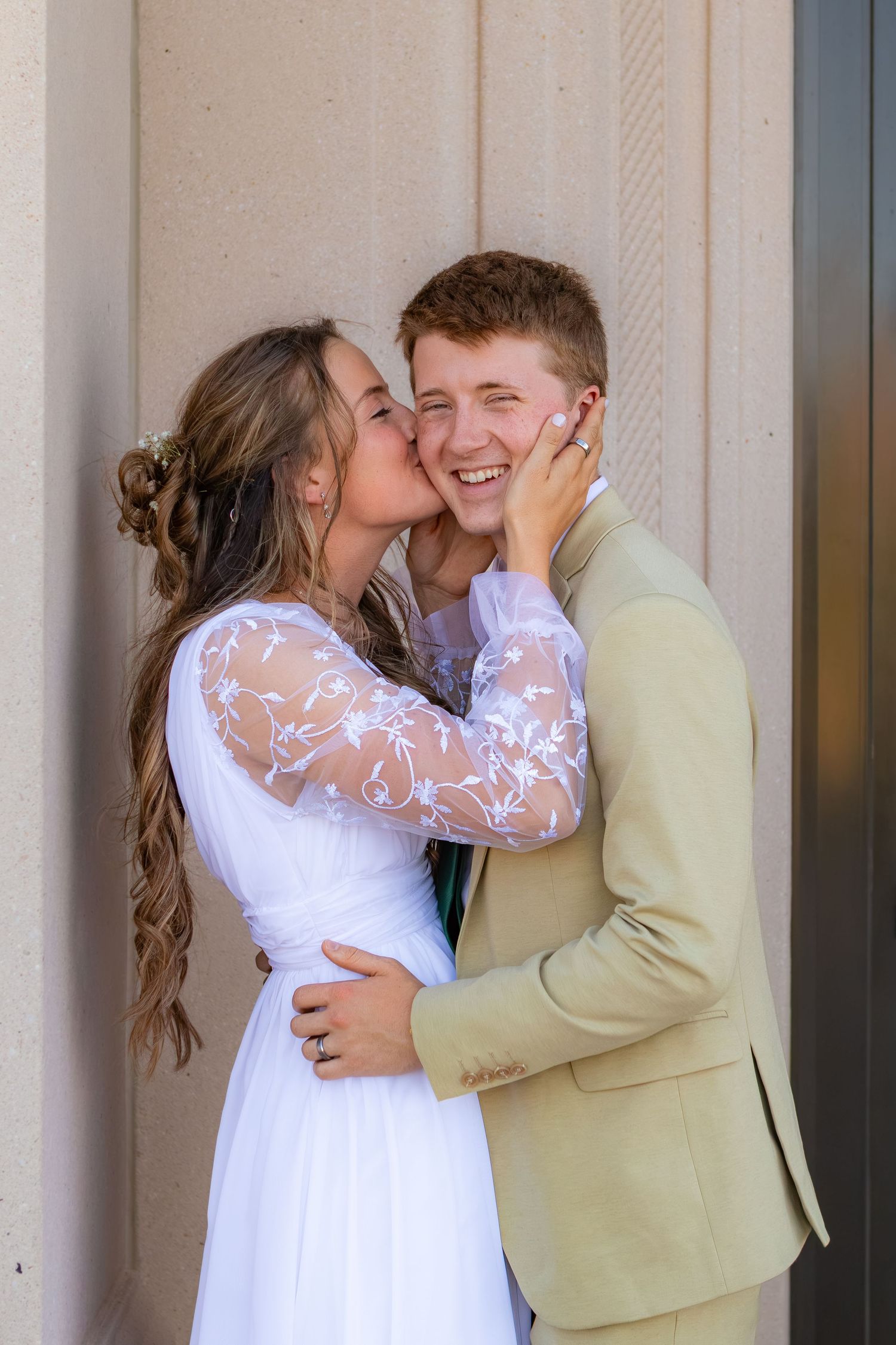 Young bride in white lace dress kisses groom in tan suit on cheek as he smiles joyfully.