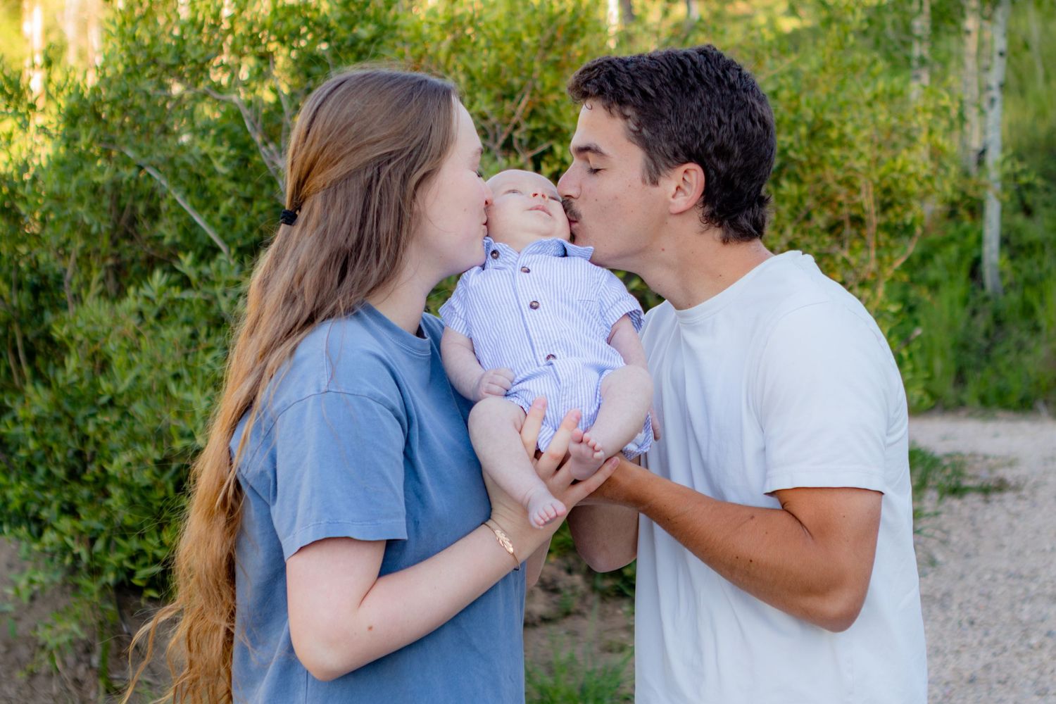 Young couple kissing their newborn baby outdoors surrounded by lush green trees on a sunny day.
