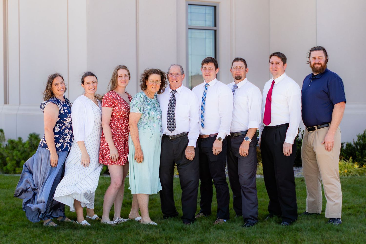 Group of nine adults posing together outdoors near a building on a sunny day, dressed in formal and semi-formal attire.