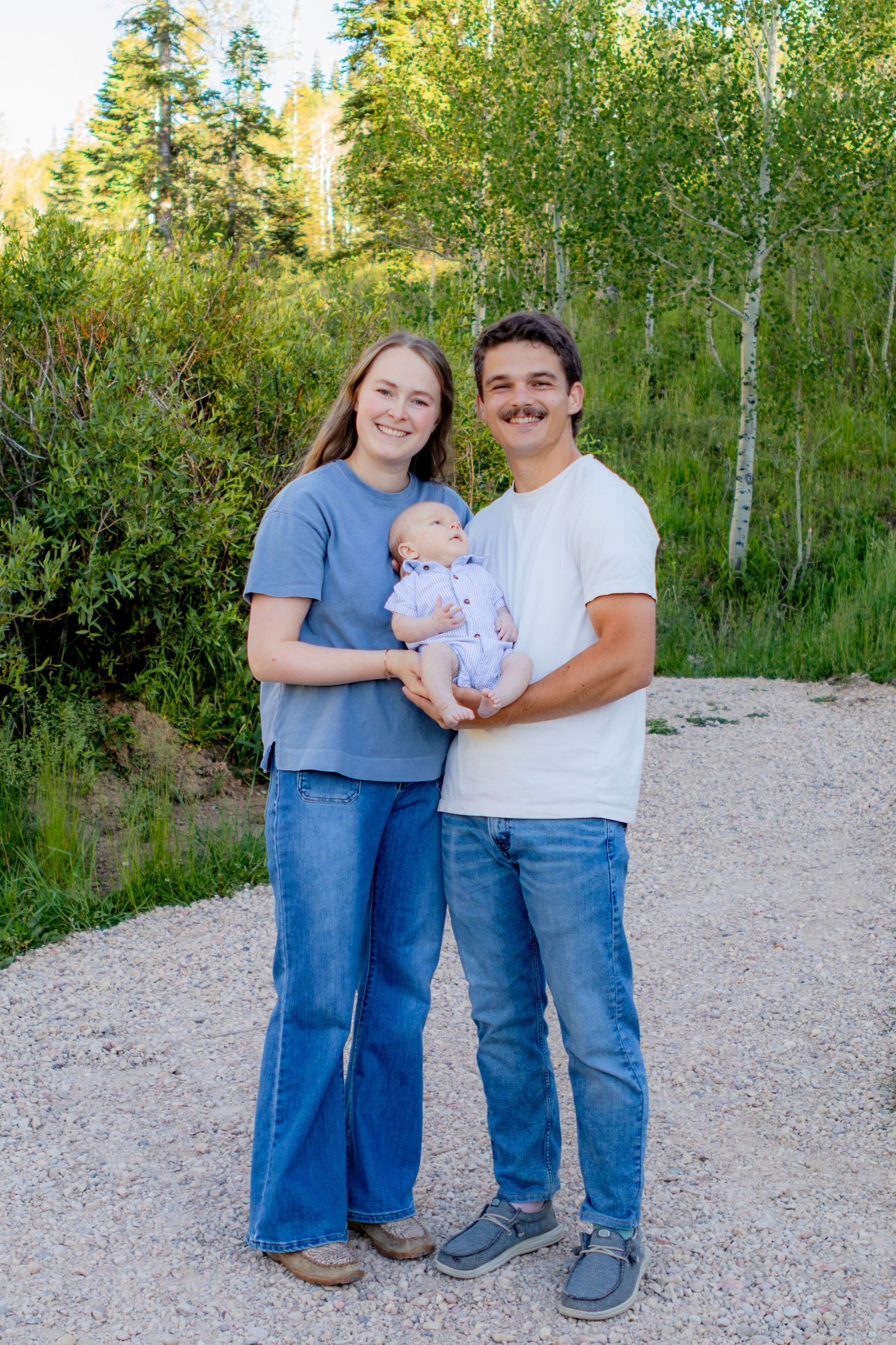 Young couple smiling outdoors holding newborn baby, standing on gravel path surrounded by green trees.