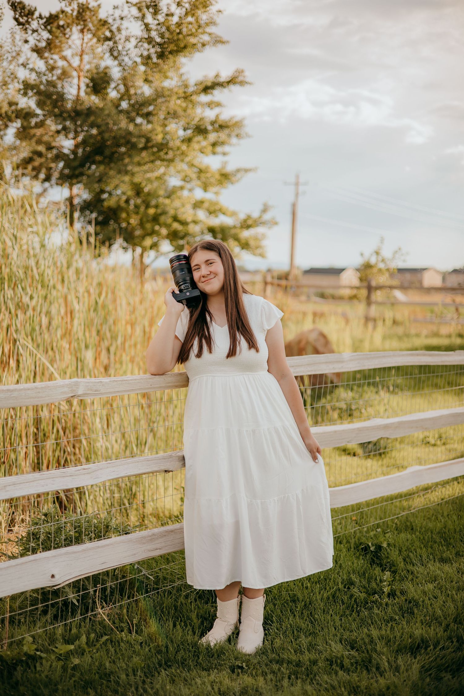 Lilly Wade Photo. Young woman in white dress holds camera while leaning on rustic wooden fence in golden hour light.