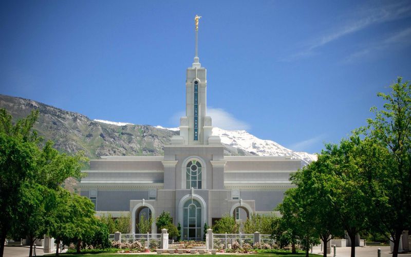 White LDS Mormon temple with tall spire against snow-capped mountains and blue sky in Utah.