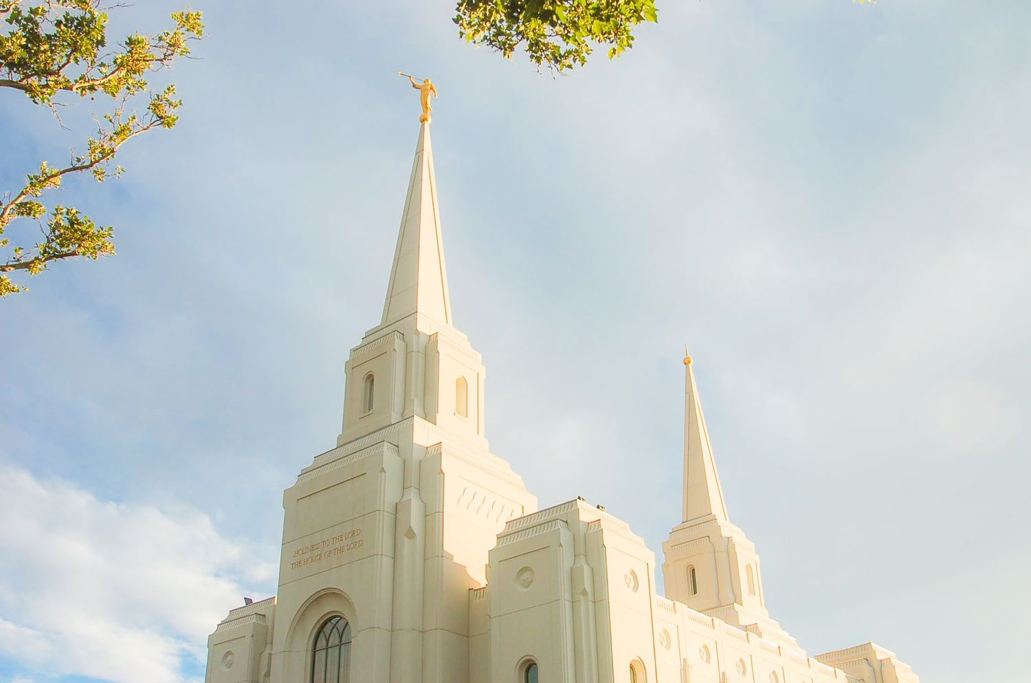 White temple with tall spires and a golden statue, lit by warm sunlight against a blue sky with soft clouds and green leaves.
