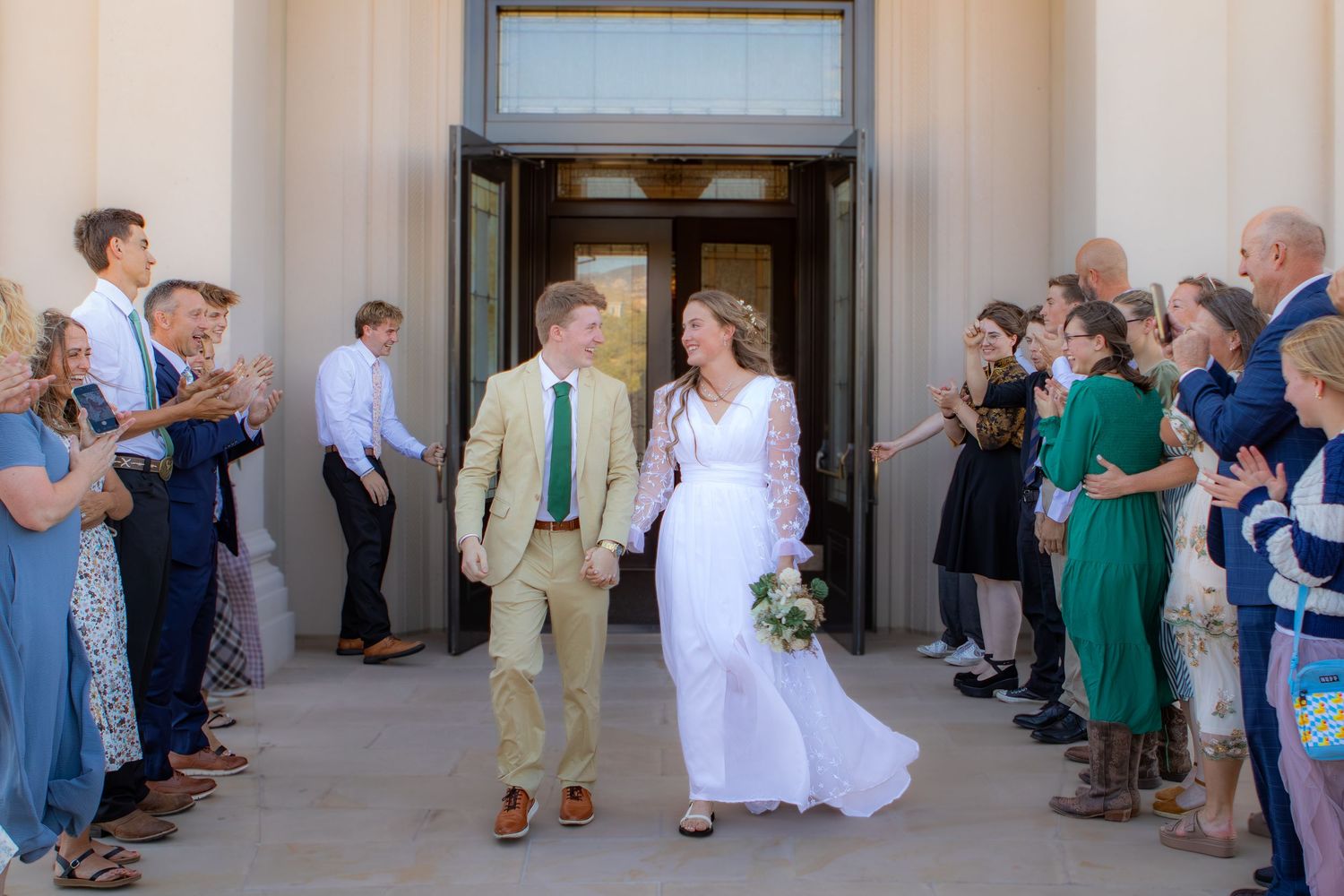 Newlywed couple walks through cheering guests outside a wedding venue, bride in white gown holding bouquet.